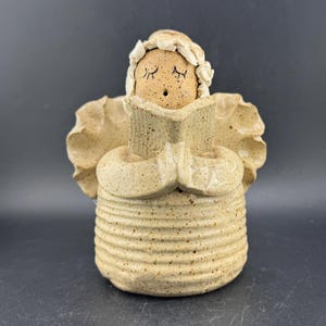 May include: A ceramic angel figurine in a beige speckled finish. The angel is reading a book, with closed eyes and a small open mouth. The figurine has large wings and a textured, ribbed body, and is wearing a head covering.