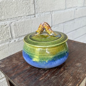 May include: A green and blue ceramic jar with a yellow and brown handle on the lid.