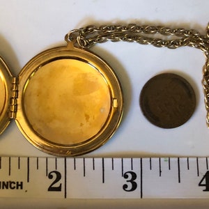 May include: Gold-toned locket necklace, open to reveal two compartments. The locket is round, with a chain. A penny is next to the locket for size comparison. The locket is approximately 1.75 inches in diameter.