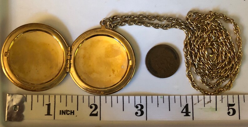 May include: Gold-toned locket necklace, open to reveal two compartments. The locket is round, with a chain. A penny is next to the locket for size comparison. The locket is approximately 1.75 inches in diameter.