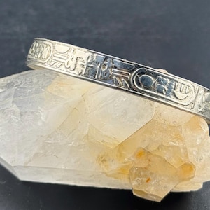 Puede incluir: Un brazalete plateado con símbolos jeroglíficos grabados, apoyado sobre un cristal transparente de color amarillo claro. El brazalete presenta grabados detallados. El fondo es de color oscuro.