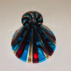May include: A decorative glass bottle with a swirl-shaped stopper. The bottle features a pattern of blue, red, and gold stripes. The bottle is approximately 10 cm tall and 7.6 cm wide.