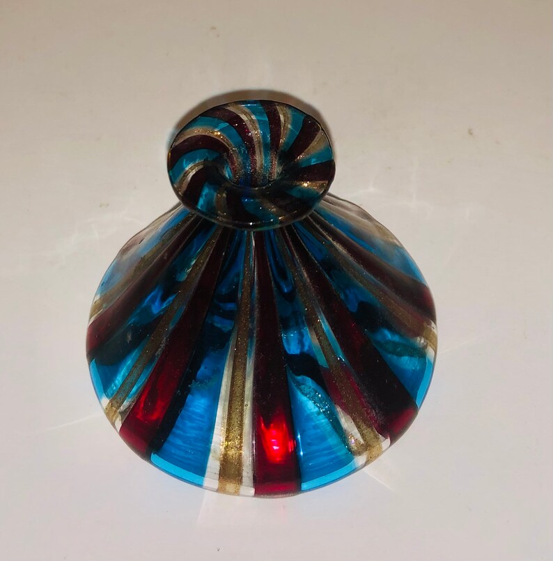 May include: A decorative glass bottle with a swirl-shaped stopper. The bottle features a pattern of blue, red, and gold stripes. The bottle is approximately 10 cm tall and 7.6 cm wide.