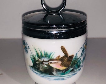 Pode incluir: Um ovo de porcelana branca com uma tampa prateada e uma alça em forma de anel. A xícara apresenta uma imagem pintada de um pássaro marrom com o peito amarelo, empoleirado em um galho verde sobre uma lagoa azul.