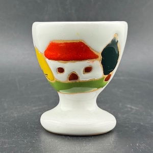 May include: White ceramic egg cup with a colorful hand-painted design. The design features a red-roofed house, green grass, and a dark green tree. The egg cup has a pedestal base and a flared top.