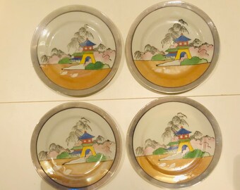 May include: Set of four white ceramic plates with a gold rim. Each plate features a hand-painted design of a Japanese pagoda with trees and flowers in shades of pink, green, blue, and yellow.