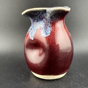 Könnte beinhalten: Eine kleine, handgefertigte Keramikvase mit einem tief roten Überzug und einem weißen und blauen Glaselement. Die Vase hat eine unregelmäßige, organische Form.