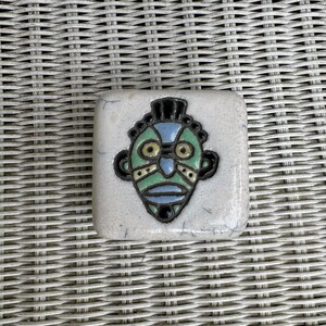 Könnte beinhalten: Eine weiße Keramikfliese mit einem bemalten Maskenmotiv in Grün, Blau und Gelb. Die Maske hat einen schwarzen Umriss und ist vor einem weißen Hintergrund platziert.