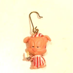 May include: A pink pig wearing red and white striped pajamas holds a candle in its right hand. The pig has a sleepy expression and is a Christmas ornament.