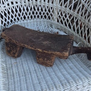 May include: A weathered, dark brown wooden footstool with a rectangular top and two short legs. The stool has a handle on one end. It is placed on a white wicker chair.