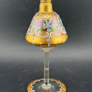 Puede incluir: Un frasco de perfume de vidrio antiguo con base y borde dorados. El frasco tiene un cuerpo acampanado en forma de campana con diseños florales y de desplazamiento en azul, rosa y blanco. La base es de vidrio transparente con un borde dorado.