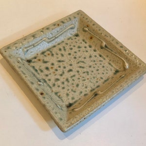 May include: Square ceramic dish with a speckled, textured surface in shades of gray and green. The rim features a raised, wavy design. This handmade dish is suitable for serving food or as a decorative accent.