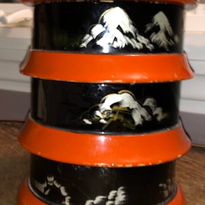 May include: A three-tiered, round, wooden box with a black base and orange trim. Each tier features a white and gold painted design of stylized clouds.