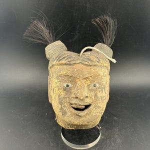 Könnte beinhalten: Eine Vintage-Holzmaske mit einem lächelnden Gesicht, mit zwei Büscheln schwarzer Haare oben. Die Maske hat grüne Augen und einen Metallring am Boden. Das Gesicht ist in Braun- und Goldtönen bemalt.
