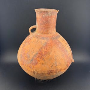 Könnte beinhalten: Eine antik anmutende Krug aus Terrakotta mit rundem Körper, schmalem Hals und kleinem Henkel. Der Krug hat ein rustikales, gealtertes Aussehen mit Farb- und Texturvariationen, was auf Handarbeit hindeutet.