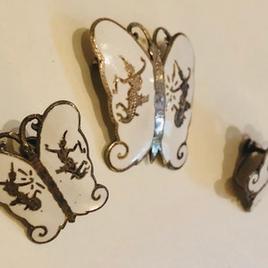 May include: Set of three silver butterfly earrings with white enamel and black detailing. The butterflies are different sizes and have a delicate design.