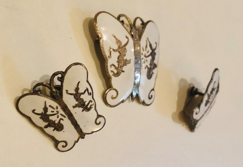 May include: Set of three silver butterfly earrings with white enamel and black detailing. The butterflies are different sizes and have a delicate design.
