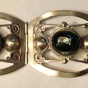 Puede incluir: Pulsera plateada con cuatro eslabones ovalados. Cada eslab&oacute;n presenta una piedra negra central con detalles iridiscentes, flanqueada por esferas plateadas y adornos en espiral. Cierre sencillo.