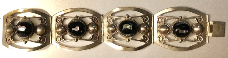 Puede incluir: Pulsera plateada con cuatro eslabones ovalados. Cada eslab&oacute;n presenta una piedra negra central con detalles iridiscentes, flanqueada por esferas plateadas y adornos en espiral. Cierre sencillo.