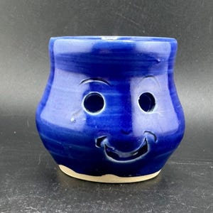 May include: A handmade, cobalt blue ceramic mug with a smiling face design. The mug features two circular eye holes and a curved mouth, giving it a playful appearance. The base is unglazed, revealing the natural clay color.