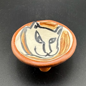 May include: A small, terracotta-colored ceramic dish with three feet. The dish is decorated with a hand-painted black and white illustration of a cat's face.