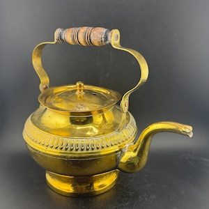 May include: A vintage brass teapot with a wooden handle. The teapot has a rounded body, a curved spout, and a lid with a small knob. The handle is attached to the body with brass brackets. The teapot is a shiny gold colour.