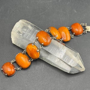 May include: A bracelet featuring seven oval-shaped, polished orange amber stones set in silver-toned metal links. The stones are connected by small metal rings, and the piece includes a rectangular metal box clasp at one end of the chain.