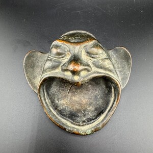 May include: A vintage bronze ashtray shaped like a face with a wide open mouth. The face has a distressed look with a green patina.