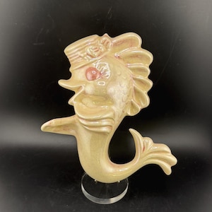 May include: A ceramic fish figurine in a light beige color, wearing a hat with a red accent on the eye. The fish has a curved tail and is mounted on a clear stand. The figurine is a decorative item.