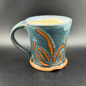 May include: A handmade ceramic mug with a teal glaze and a brown design of stylized leaves. The mug has a wide base and a rounded handle.