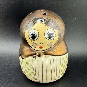 May include: A ceramic doll-shaped salt and pepper shaker. The top is bronze-coloured, and the face is light yellow with large blue eyes and a red mouth. The bottom half is white with vertical black stripes and green polka dots.