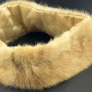 May include: A vintage fur collar in a light tan colour. The fur is thick and plush, with a soft, luxurious texture. The interior lining is a light cream-coloured satin. The collar is designed to be worn around the neck.