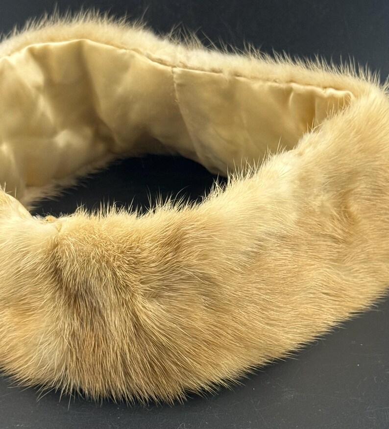 May include: A vintage fur collar in a light tan colour. The fur is thick and plush, with a soft, luxurious texture. The interior lining is a light cream-coloured satin. The collar is designed to be worn around the neck.