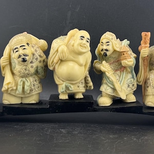 Peut inclure: Ensemble de sept figurines ivoire représentant des divinités japonaises traditionnelles. Chaque figurine est finement détaillée, avec des poses et des accessoires variés, montées sur une base noire. Les figurines présentent diverses expressions et tenues.