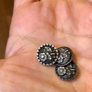 May include: Three silver-toned buttons with a floral design. The buttons are round and have a raised edge.