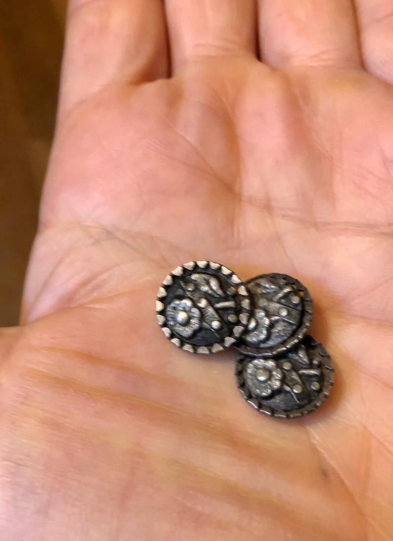 May include: Three silver-toned buttons with a floral design. The buttons are round and have a raised edge.