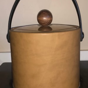 May include: A brown leather ice bucket with a black leather handle and a wooden knob on the lid. The bucket has a gold and brown trim around the bottom.