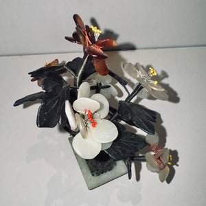 May include: A decorative floral arrangement featuring white, brown, and translucent flowers with yellow stamens and red accents. The flowers are set in a square, green stone base with dark green leaves.