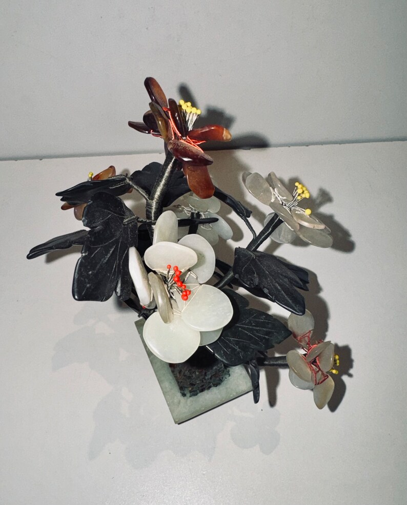 May include: A decorative floral arrangement featuring white, brown, and translucent flowers with yellow stamens and red accents. The flowers are set in a square, green stone base with dark green leaves.