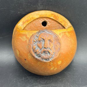 May include: A round, ochre-colored ceramic wall pocket featuring a textured surface and a central circular medallion depicting a bearded male face in profile. The vessel has an open top with a small hole for mounting, showcasing a rustic finish.