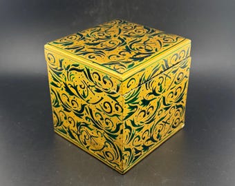 Könnte beinhalten: Quadratische dekorative Box mit Scharnierdeckel. Die Box ist goldfarben lackiert und mit einem dunkelgrünen, wirbelnden Blumenmuster versehen. Die Box steht auf einer schwarzen Oberfläche.