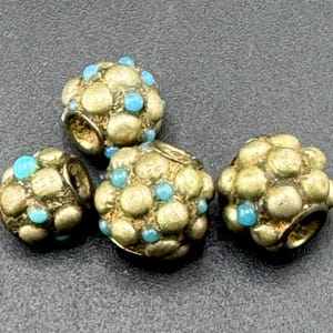 May include: Four antique, round beads with a textured, gold-colored surface. Each bead is adorned with small, turquoise-colored accents. The beads have a hole for stringing, suggesting they are for jewelry making or similar crafts.