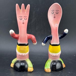 May include: A whimsical pair of ceramic salt and pepper shakers, one a fork and the other a spoon. Both are pink with surprised faces, arms, and legs. The fork has a red arm, the spoon a blue arm. They have yellow, green, and black accents.
