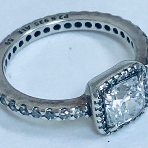 May include: A silver ring with a square-shaped clear stone set in a silver frame. The ring is decorated with small clear stones around the band. The ring is marked "925 ALE 90".