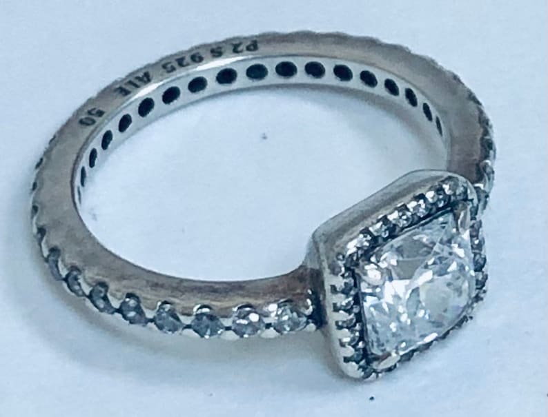May include: A silver ring with a square-shaped clear stone set in a silver frame. The ring is decorated with small clear stones around the band. The ring is marked "925 ALE 90".