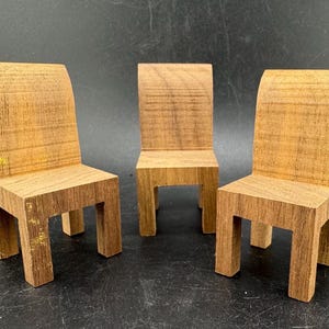 May include: Three miniature wooden chairs with a natural wood grain finish. Each chair has a rectangular seat and legs, with a curved backrest. The chairs are arranged in a row against a dark background.