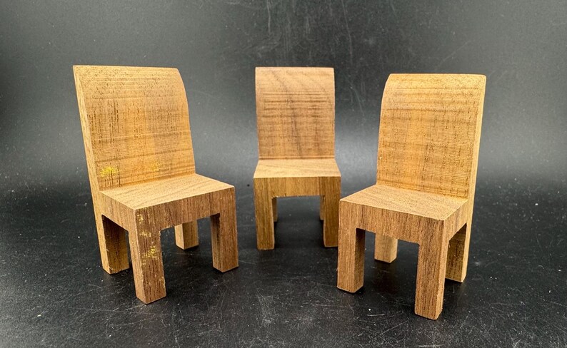 May include: Three miniature wooden chairs with a natural wood grain finish. Each chair has a rectangular seat and legs, with a curved backrest. The chairs are arranged in a row against a dark background.