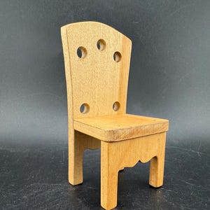 May include: A small, wooden chair with a light brown finish. The chair has a rectangular seat and a back with four circular cutouts. The legs are straight, and the back has a curved top. The chair is approximately 6 inches tall.