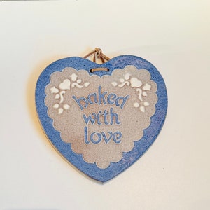 Può includere: Un cartello in ceramica a forma di cuore blu e bianco con il testo "baked with love" in lettere bianche.