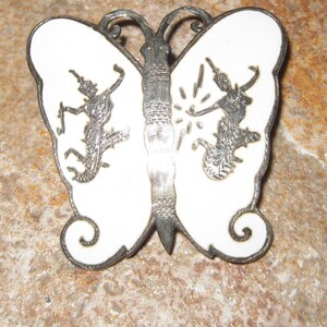 May include: A set of three silver butterfly brooches with white enamel. Each brooch features a stylized butterfly with intricate details and silhouettes of figures.